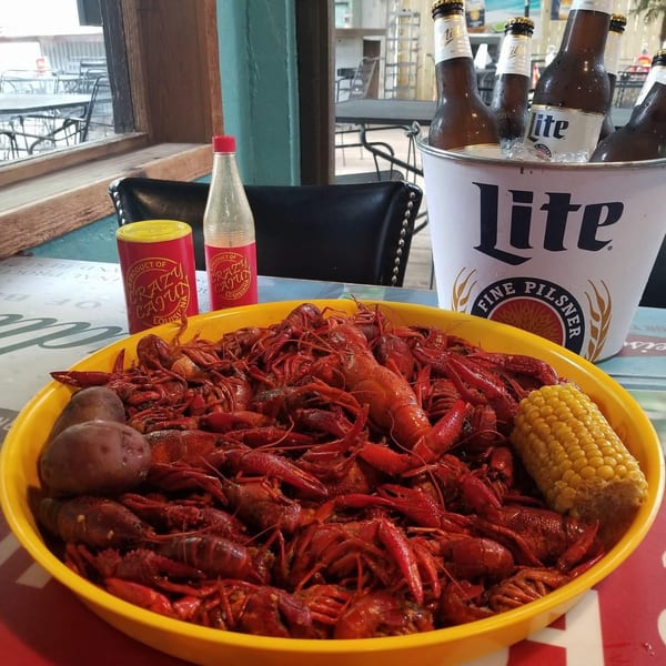 A large yellow bowl of boiled crawfish, corn, and potatoes with beer and seasoning on a table.