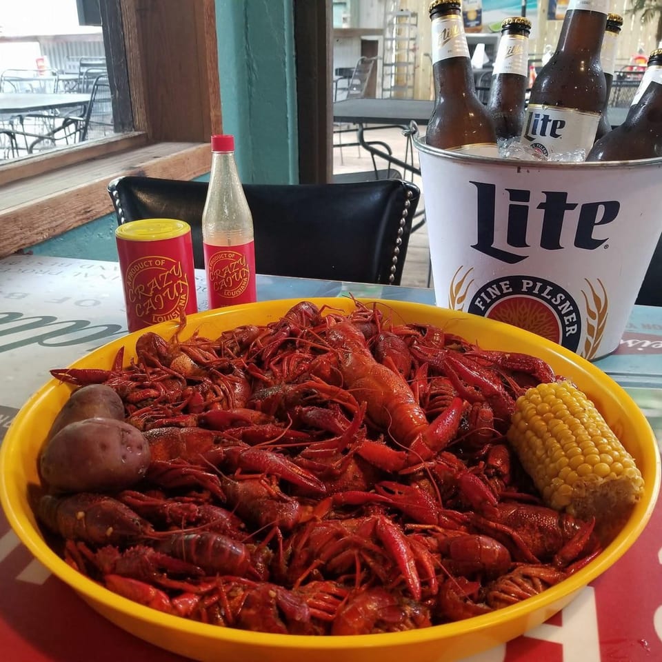 A large yellow bowl of boiled crawfish, corn, and potatoes with beer and seasoning on a table.